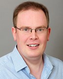 A photo of Ian Crawford against a grey background. He has short brown hair, blue eyes and is wearing glasses. He is wearing a pale blue shirt and is smiling at the camera.