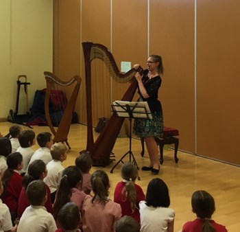 A photo of harpist Natasha Gale performing in front of the pupils, who are sat on the floor watching her